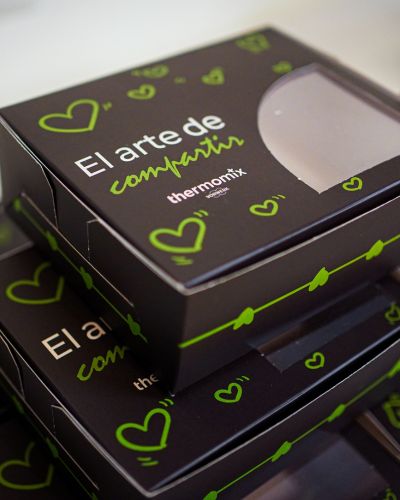 Cajas negras con corazones verdes y el texto “El arte de compartir” de Thermomix, apiladas como empaque de regalo.