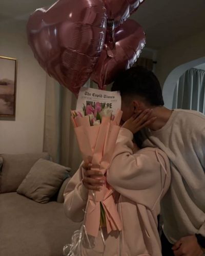 Momento en pareja en el Día de San Valentín. Él le da un beso en la mejilla y ella sostiene unos tulipanes y unos globos en forma de corazón 