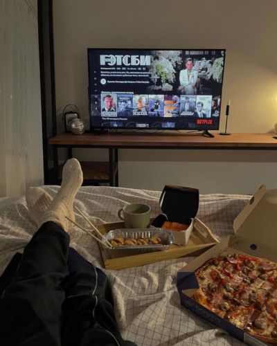 Persona recostada en cama viendo Netflix por la noche con pizza y snacks en una bandeja.