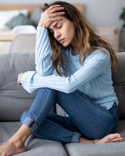 Mujer sentada en un sillón con gesto de dolor y mano en la frente, reflejando malestar físico y agotamiento por mastitis.
