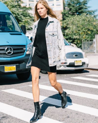 Mujer caminando con  mezclilla claro sobre vestido negro corto y botas negras, look urbano moderno.