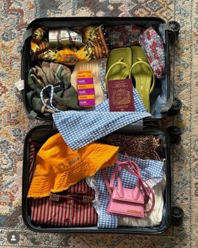 Maleta abierta con ropa, accesorios, sandalias, sombrero, pasaporte y artículos de viaje acomodados en su interior.