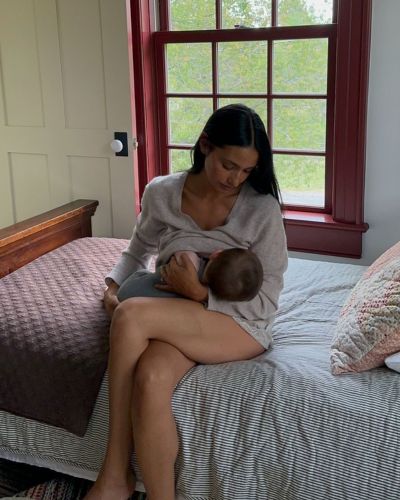 Mujer en periodo de lactancia amamantando a su bebé sentada en la cama junto a una ventana.
