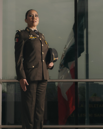 Mujer en uniforme militar de la Secretaría de la Defensa Nacional posando junto a ventanales, con gorra en mano; imagen para nota sobre General Ma. Dolores Domínguez Rocha y Colegio Militar.