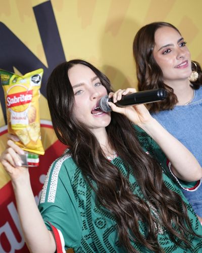 Belinda con micrófono levantando una bolsa de papas fritas en un evento con fondo de marca