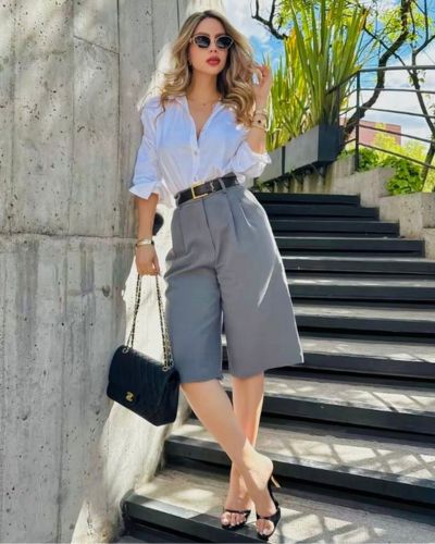 Modelo viste bermudas sastre de mujer en color gris junto a una camisa blanca y accesorios en color negro.