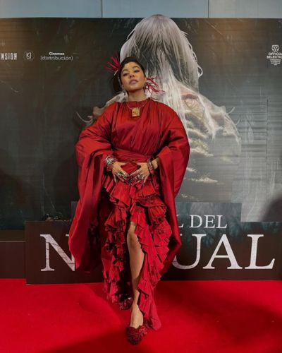 Alejandra Herrera posa en alfombra roja con un vestido rojo de capas frente a un fondo promocional de “El ritual del nahual”.