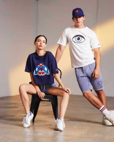 Dos modelos posan en estudio usando outfits casuales: ella con playera azul marino y shorts, él con playera blanca estampada y shorts, ambos con gorra.