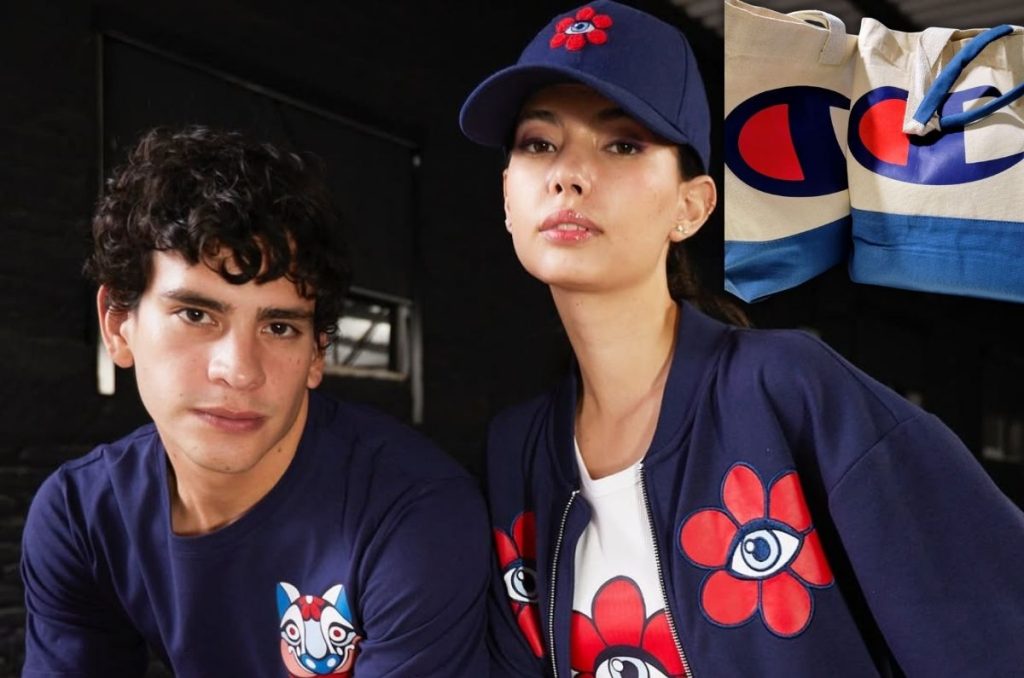 Dos modelos posan en primer plano con prendas de la colección Champion X Sofía Castellanos: él con playera azul marino con gráfico y ella con gorra y chamarra azul con flores rojas y ojos; al fondo se ve una tote bag de Champion.