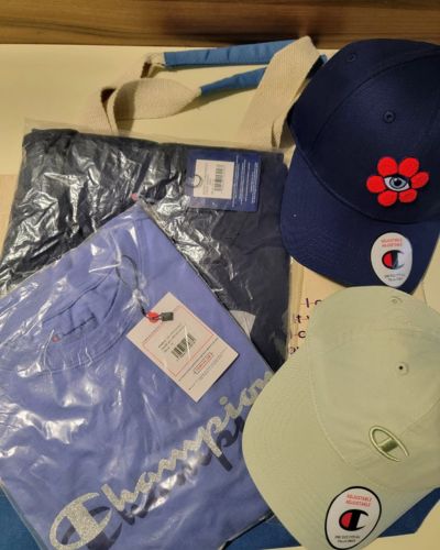 Prendas y accesorios Champion con ilustraciones coloridas, incluyendo dos gorras y una tote bag, acomodados sobre una superficie.