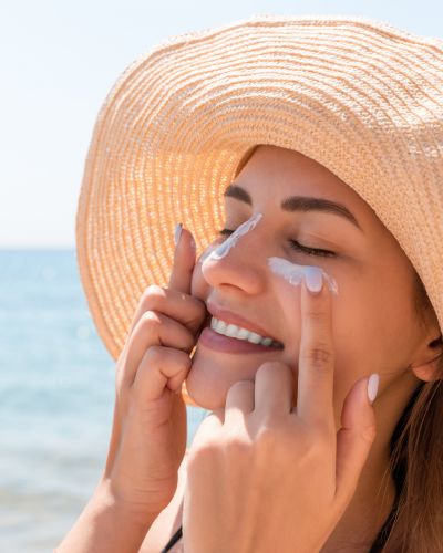 Mujer en la playa con sombrero aplicando protector solar en el rostro, cuidando la piel para mantenerla hidratada y luminosa.