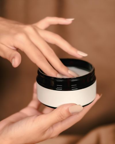 Mano tomando crema de un tarro cosmético, paso de hidratación para mantener la piel suave.