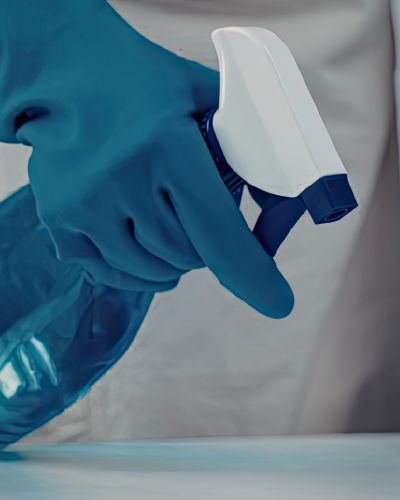 Mano con guante azul sosteniendo un atomizador de limpieza sobre una superficie, relacionado con el uso de guantes para el hogar.