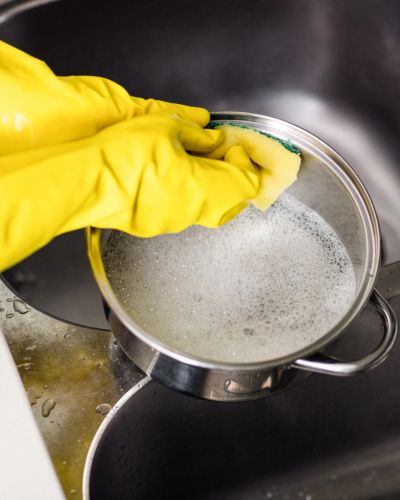 Mano con guante amarillo lavando una olla con esponja en el fregadero, ejemplo de uso de guantes para el hogar al lavar trastes.