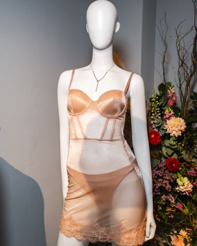 Maniquí con vestido lencero satinado en tono nude con encaje, exhibido junto a una decoración floral dentro de Guess House.