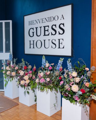 Letrero “Bienvenido a GUESS HOUSE” sobre pared azul, con bases blancas llenas de arreglos florales en la entrada de la experiencia.