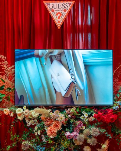 Instalación con cortinas rojas, arreglos florales y una pantalla con contenido de la marca, con el emblema triangular de Guess México al fondo en Guess House.
