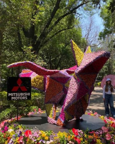 Escultura floral multicolor de gran tamaño con estilo geométrico tipo alebrije, colocada en un jardín, con el logotipo de Mitsubishi Motors México a un lado.