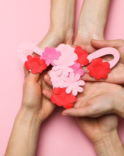 Manos sosteniendo una figura de útero hecha de papel en tonos rosa y rojo sobre fondo rosa, símbolo de salud menstrual y ciclo menstrual.