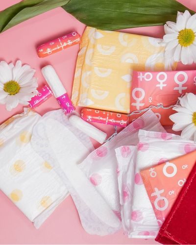 Productos de higiene menstrual y cuidado íntimo sobre fondo rosa, con toallas, tampones y empaques coloridos, relacionados con un kit de cuidado íntimo.