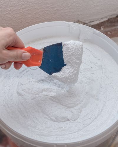 Mano mezclando una pasta blanca en una cubeta con una espátula, preparación de impermeabilizante casero para sellar superficies.