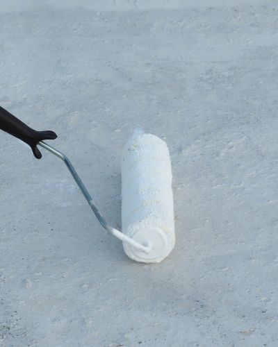 Rodillo aplicando una capa blanca sobre una superficie gris, proceso de impermeabilizar el techo para crear una barrera contra el agua.