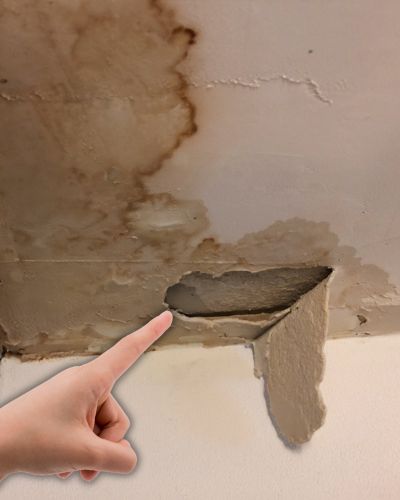 Techo interior con manchas grandes de agua y un pedazo de plafón desprendido; una mano señala el daño por filtración.