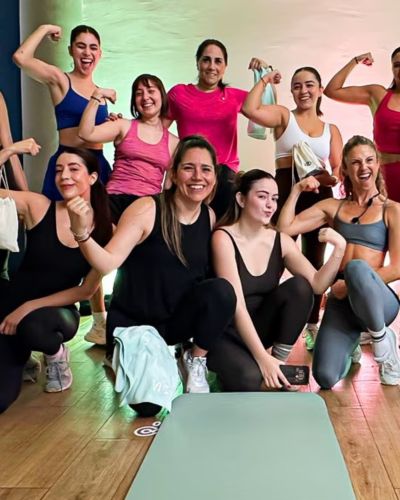 Grupo de mujeres en ropa deportiva posando juntas y flexionando los brazos, en un ambiente tipo clase fitness, como parte de una dinámica de bienestar de Vita por Lala.