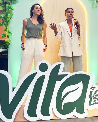 Dos mujeres en un evento de Vita por Lala frente a un gran logo “Vita”, presentando la nueva etapa de Vita México enfocada en bienestar.