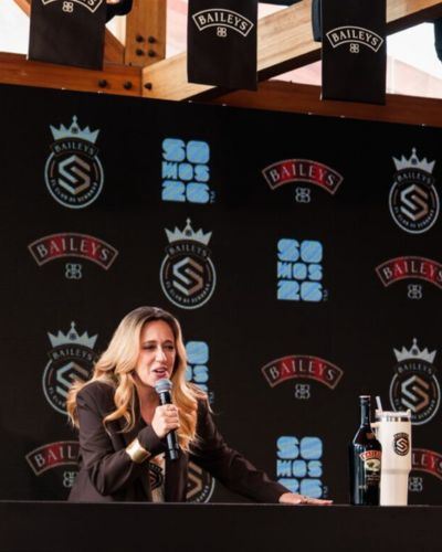 Mariana Treviño habla con micrófono durante un evento de Baileys, frente a un fondo con logos del Club de Señoras de Baileys, junto a una botella y una bebida con Baileys.