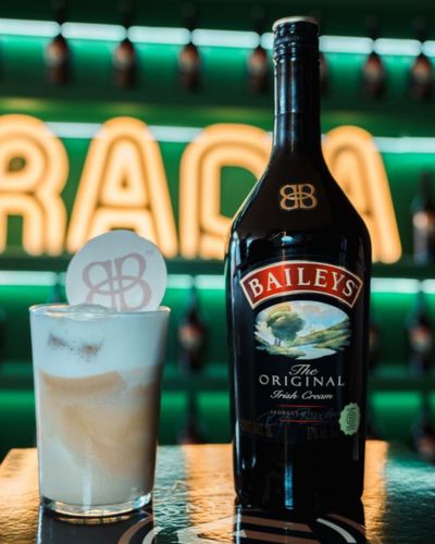 Botella de Baileys The Original Irish Cream junto a un vaso con café con Baileys servido con espuma, sobre una barra con luces verdes y branding del Club de Señoras.